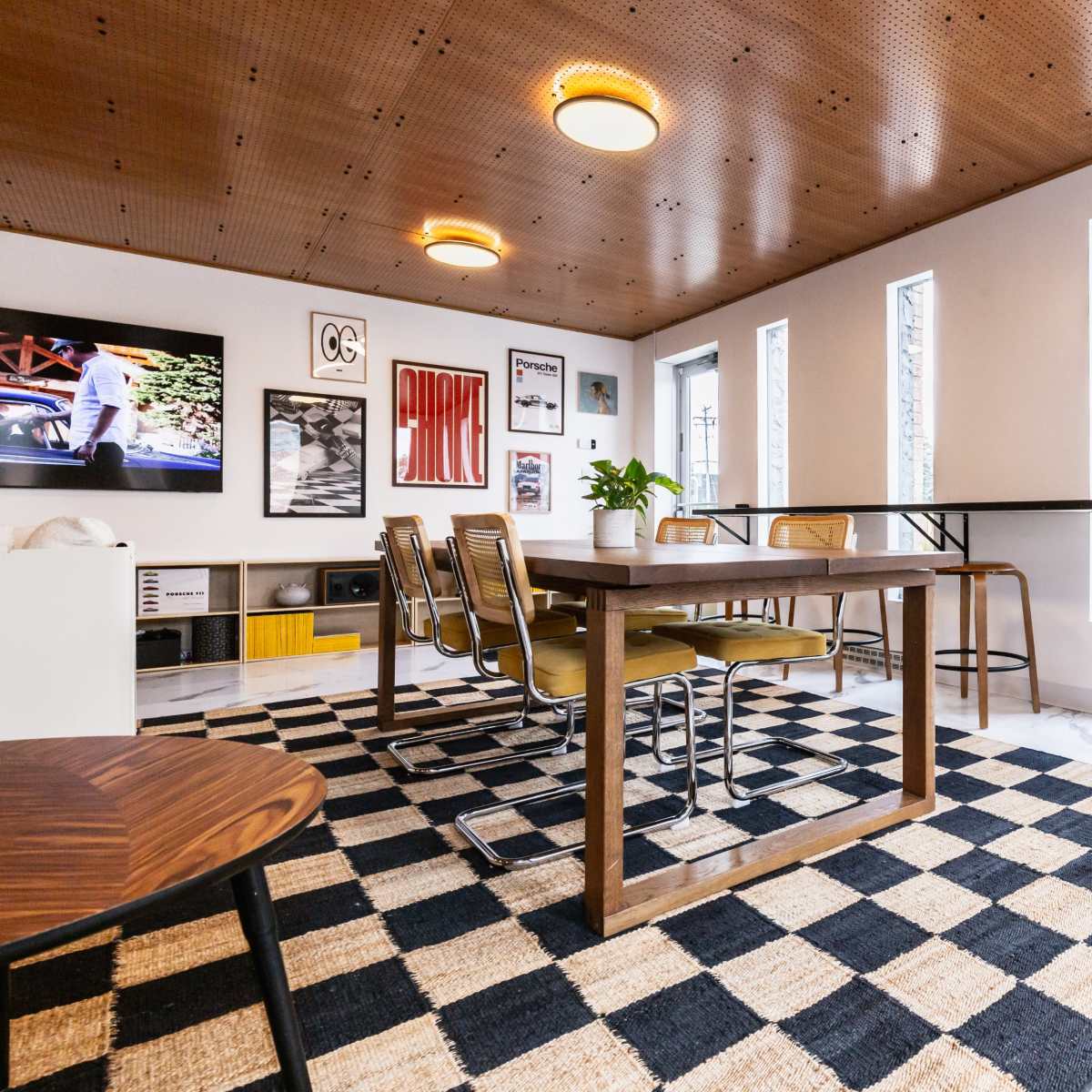 A modern dining area with a large wooden table surrounded by metal and wood chairs, set on a checkered rug, with plants and artwork on the walls.