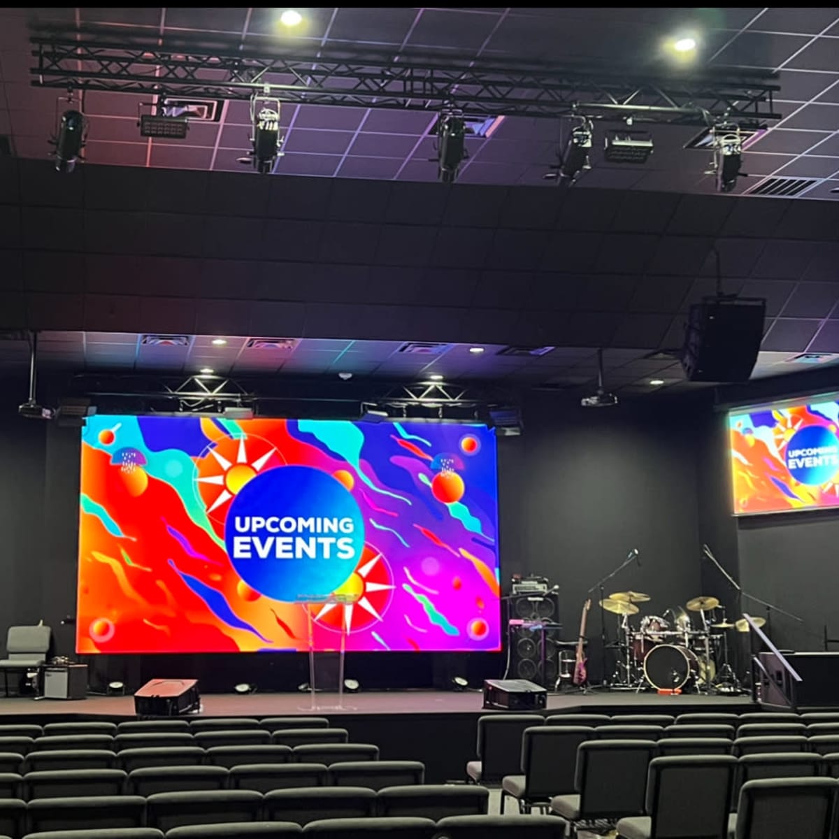 An indoor stage setup for a venue, featuring a large screen displaying "UPCOMING EVENTS" and a drum set and musical instruments arranged on the sides.