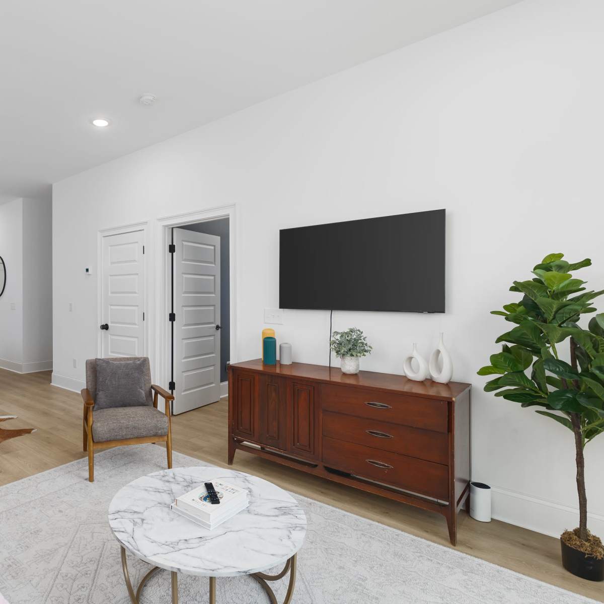 A modern living room with a seating area, a television mounted on the wall, a wooden sideboard, and an adjoining kitchen visible in the background.