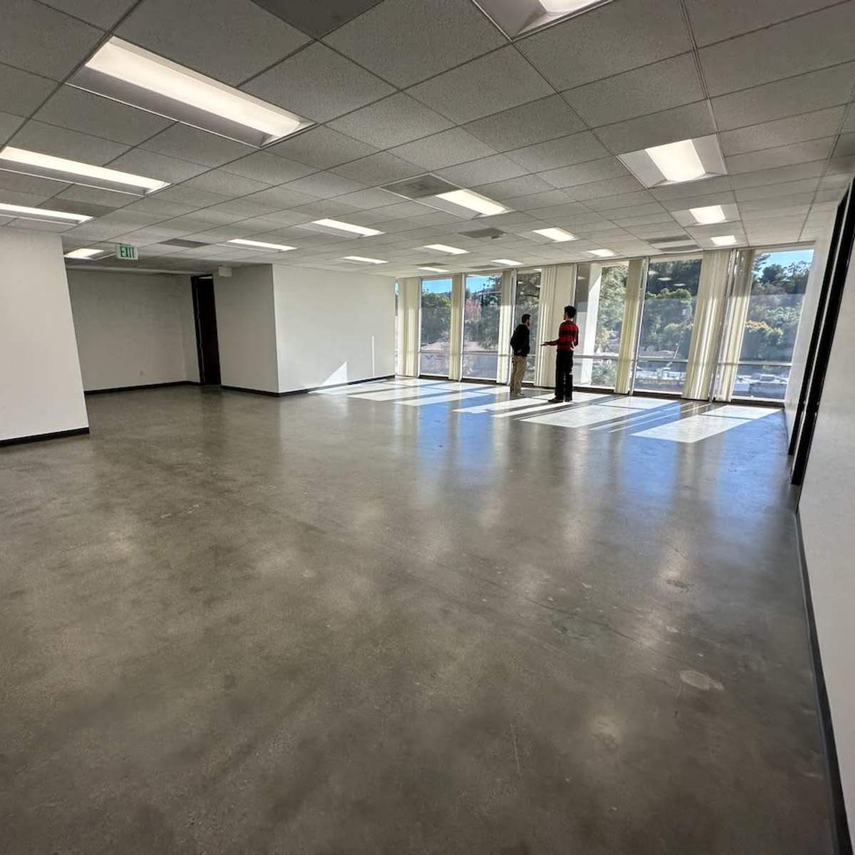 An empty room with large windows casts shadows onto a concrete floor, while two people stand near a partitioned area.
