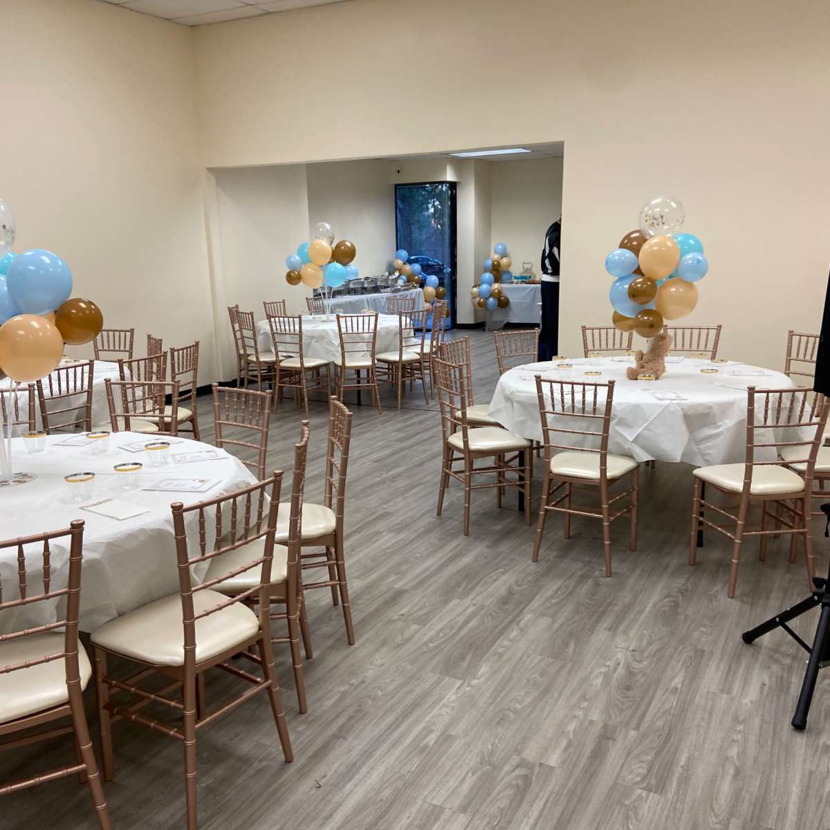 A hall with several round tables covered in white tablecloths, each surrounded by wooden chairs, and decorated with balloons in blue, brown, and clear colors.