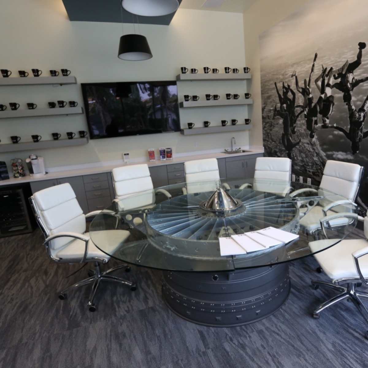 A modern conference room features a glass circular table surrounded by white leather chairs, with a wall mural of people skydiving and shelves displaying coffee mugs.