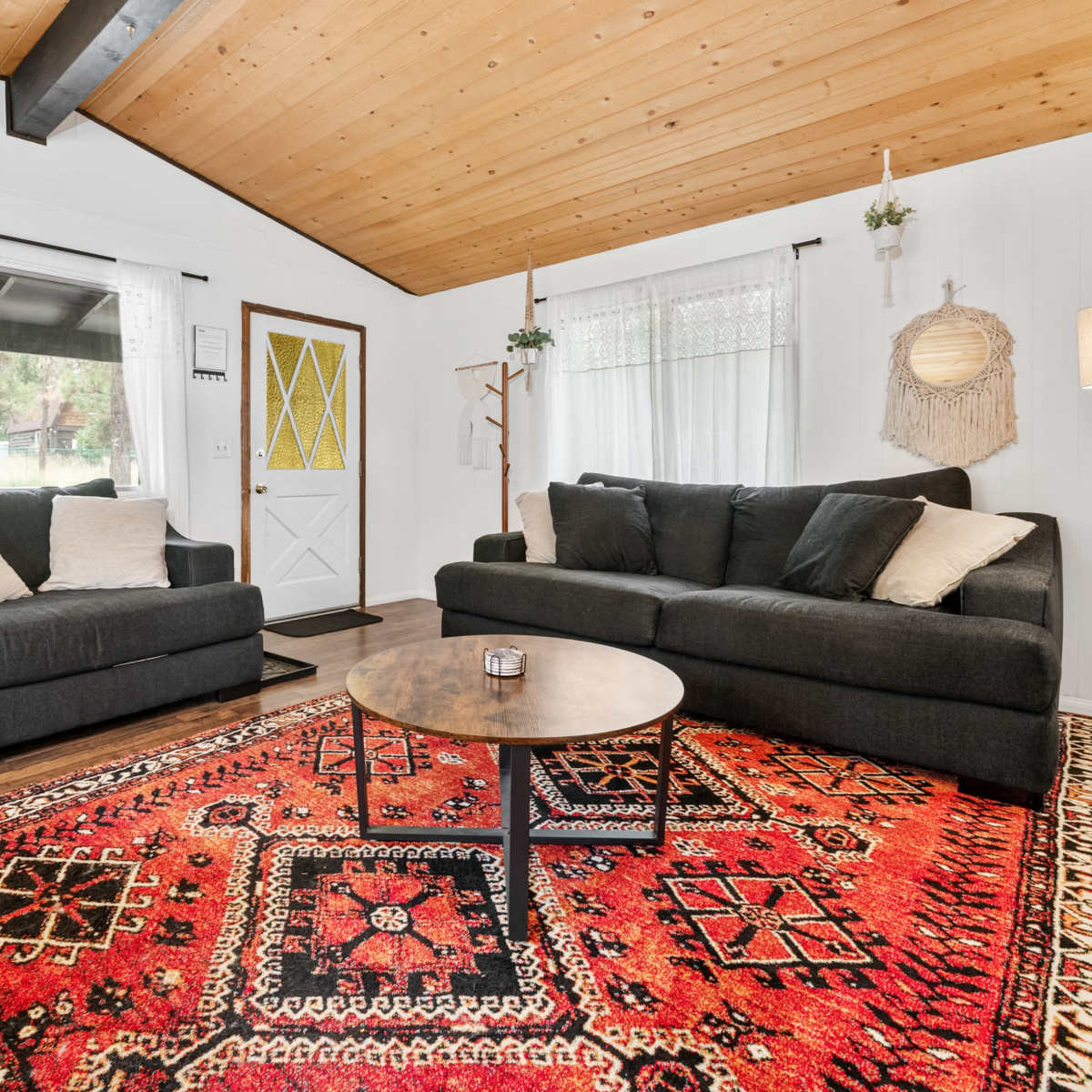 The living room features two dark sofas facing a wooden coffee table on a patterned red area rug, with a front door and large windows providing natural light.