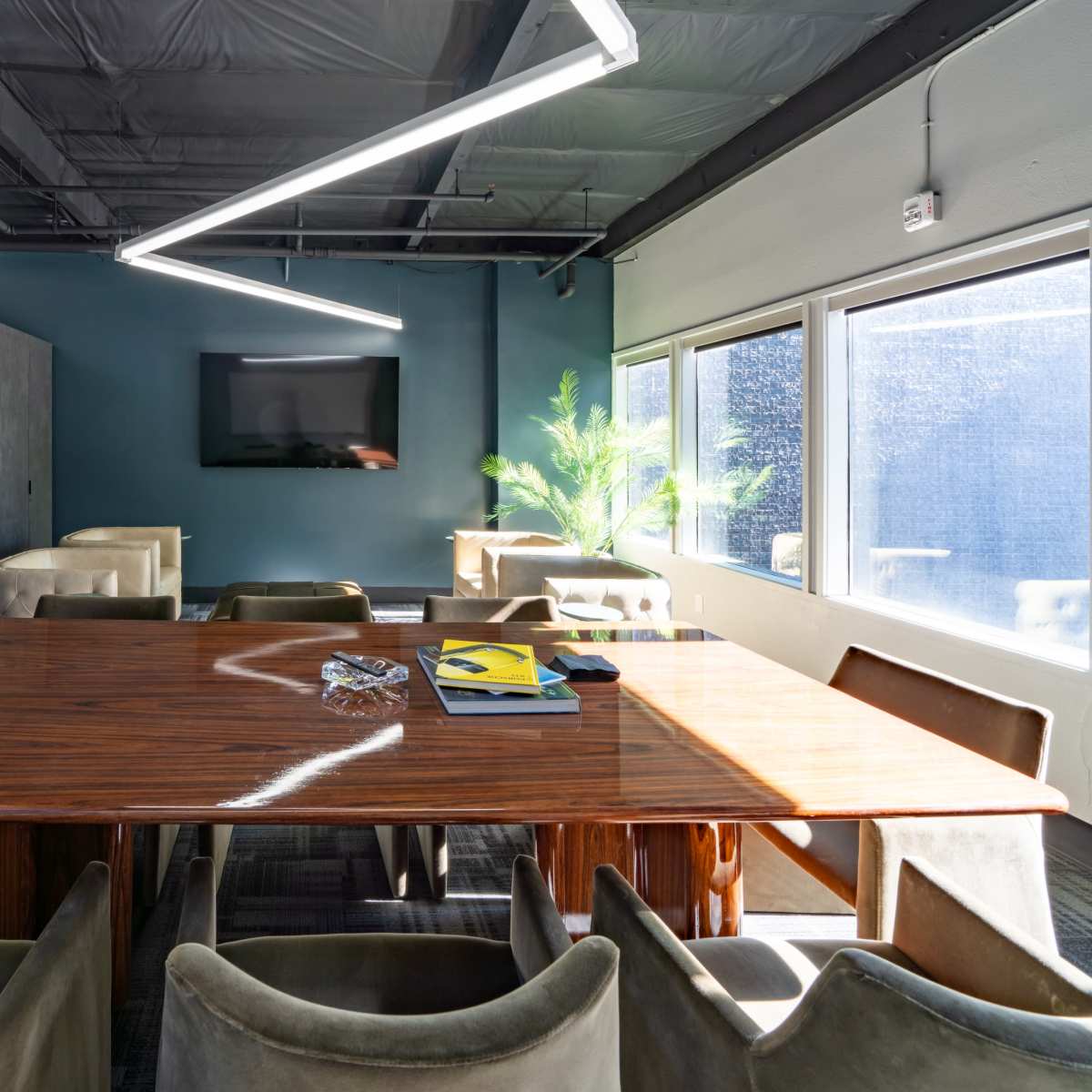 A modern conference room features a large wooden table surrounded by plush chairs, with a television mounted on the wall and natural light streaming through the windows.