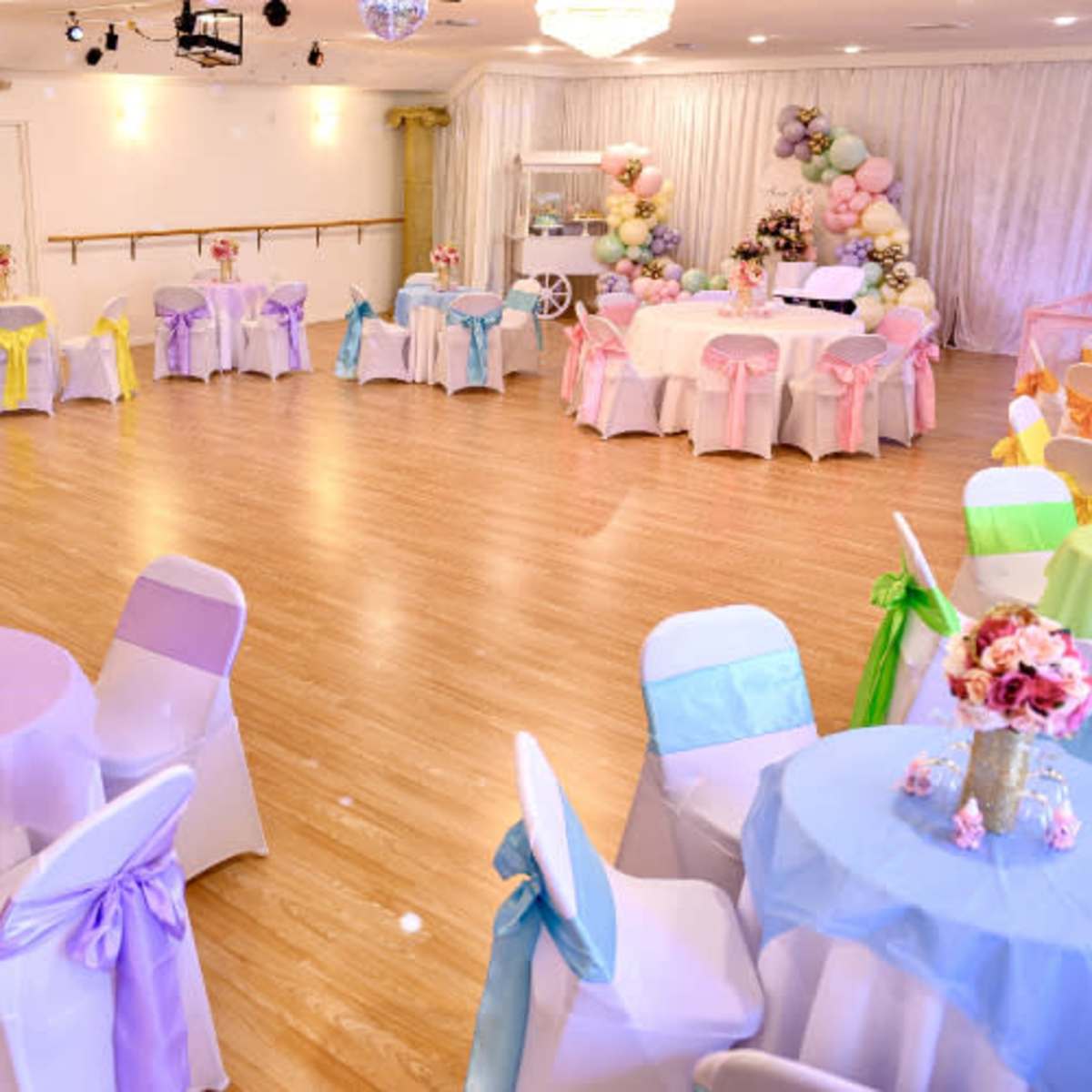 A decorated event space features round tables with colorful tablecloths and sashes, surrounded by chairs, and adorned with floral centerpieces.
