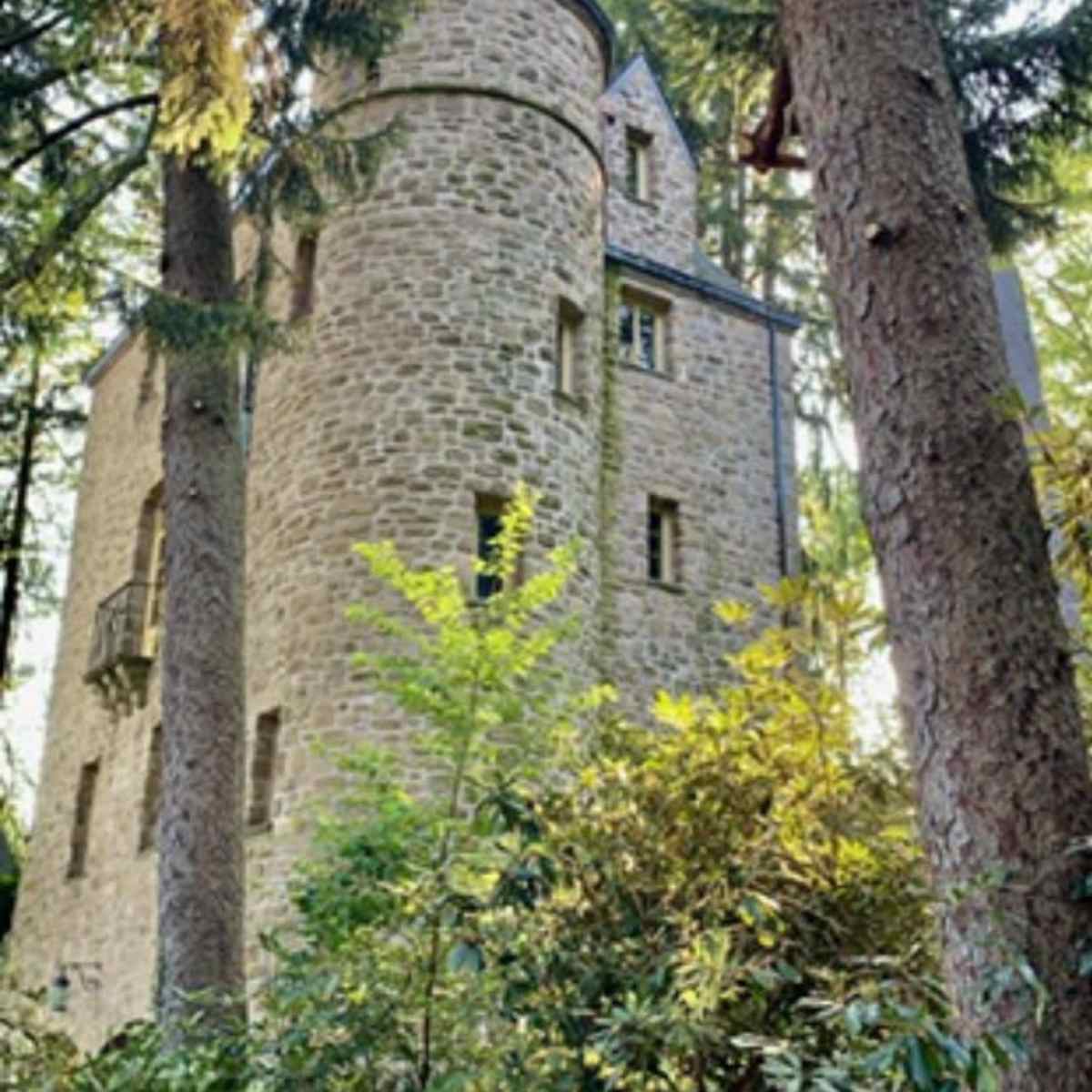 A stone tower with several windows and a balcony is nestled among tall trees and dense foliage.