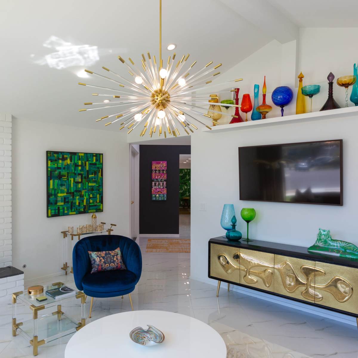 A modern living room features a gold starburst chandelier, colorful glass vases on a shelf, and a blue velvet chair.