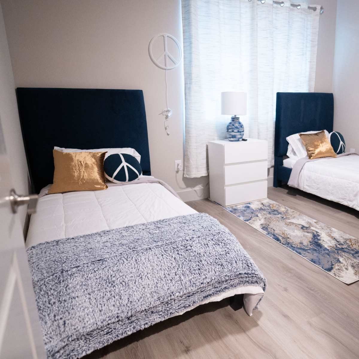 A spacious bedroom with two twin beds, a white dresser, and a window with sheer curtains, featuring a peaceful blue and white color scheme.