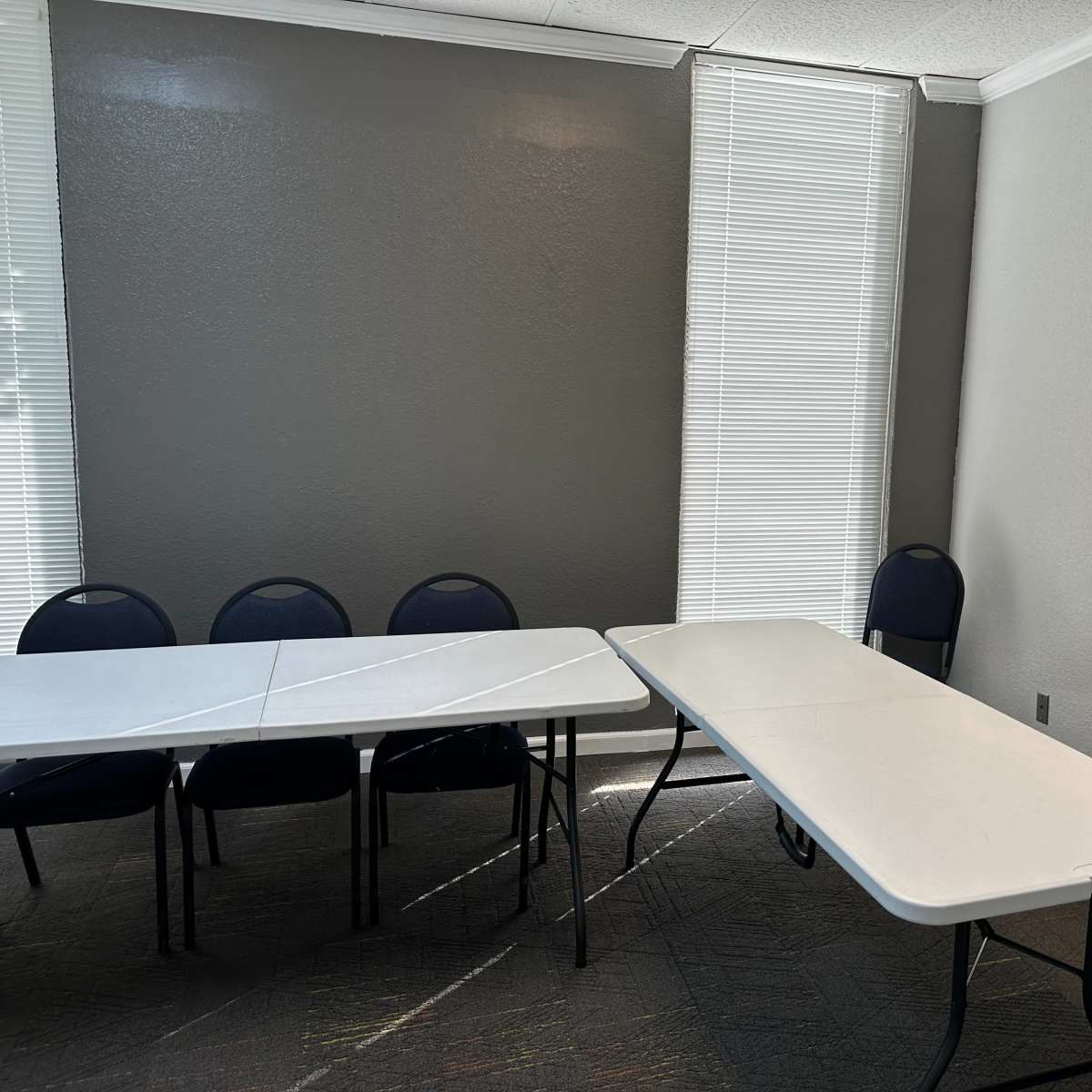 A small meeting room features two folding tables and several chairs arranged along the walls, with light streaming through window blinds.