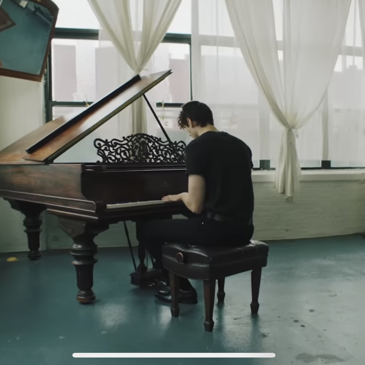 A person sits at a grand piano in a spacious, sunlit room with sheer curtains and an artwork mounted on the wall.