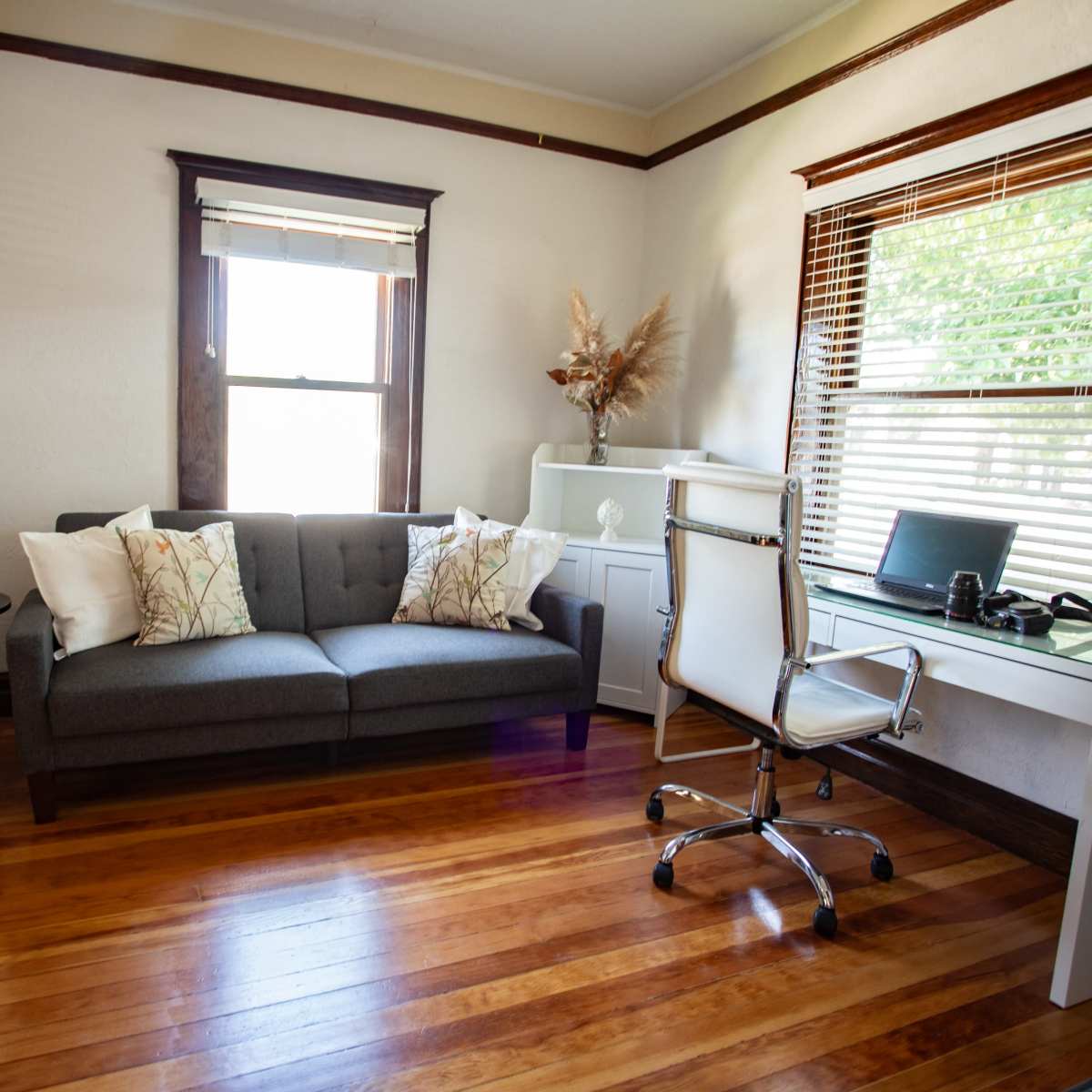 A modern office space with a gray couch, a round black side table, a white desk with a computer, and large windows allowing natural light to enter.