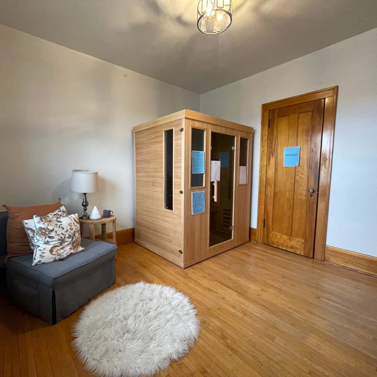 The room features a wooden sauna in one corner, a gray chair with a decorative pillow, and a small round rug on the hardwood floor.