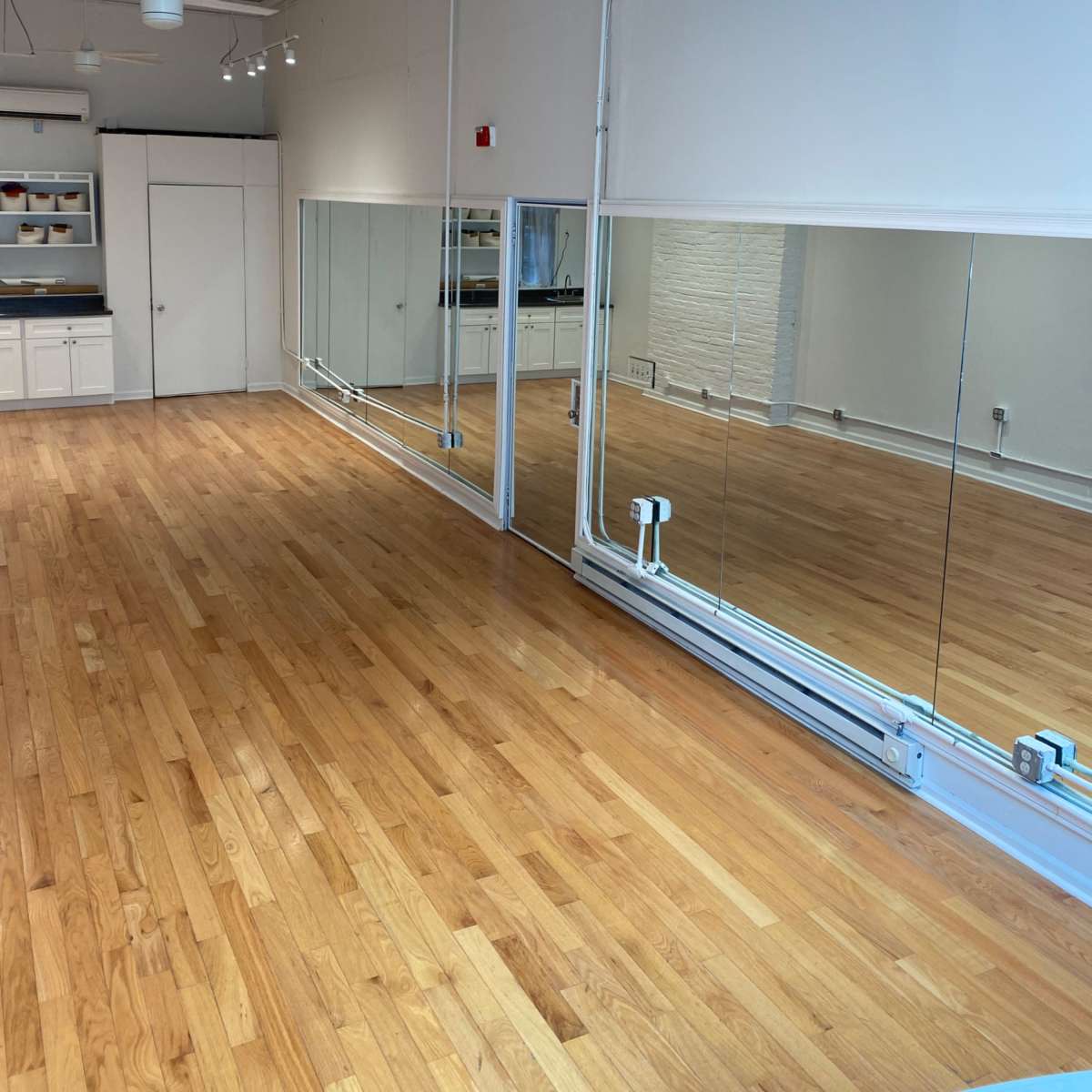 A spacious, well-lit room with wooden floors, large mirrors on one wall, and overhead lighting fixtures.