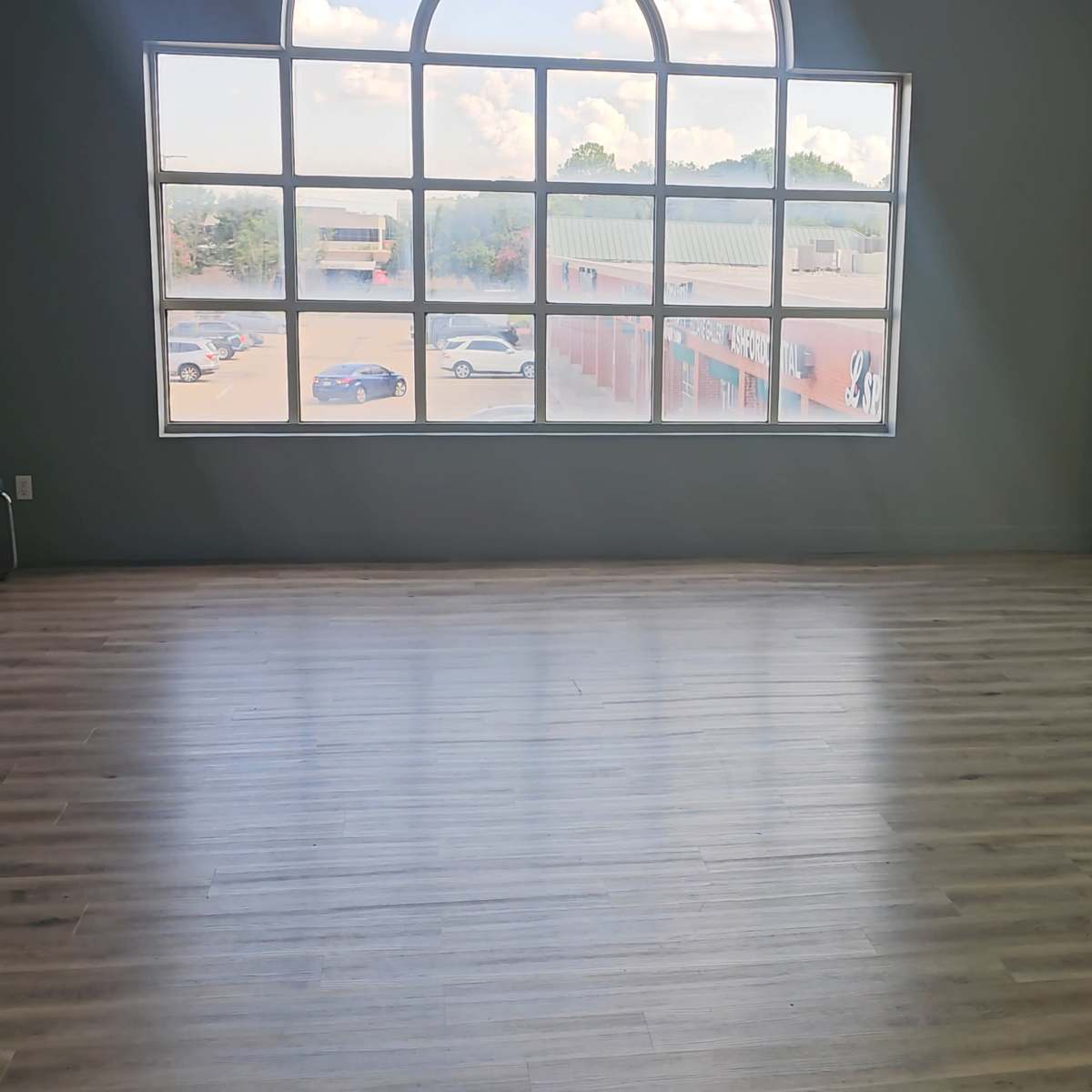 A spacious room with a wooden floor and large arched windows letting in natural light overlooks a parking lot.