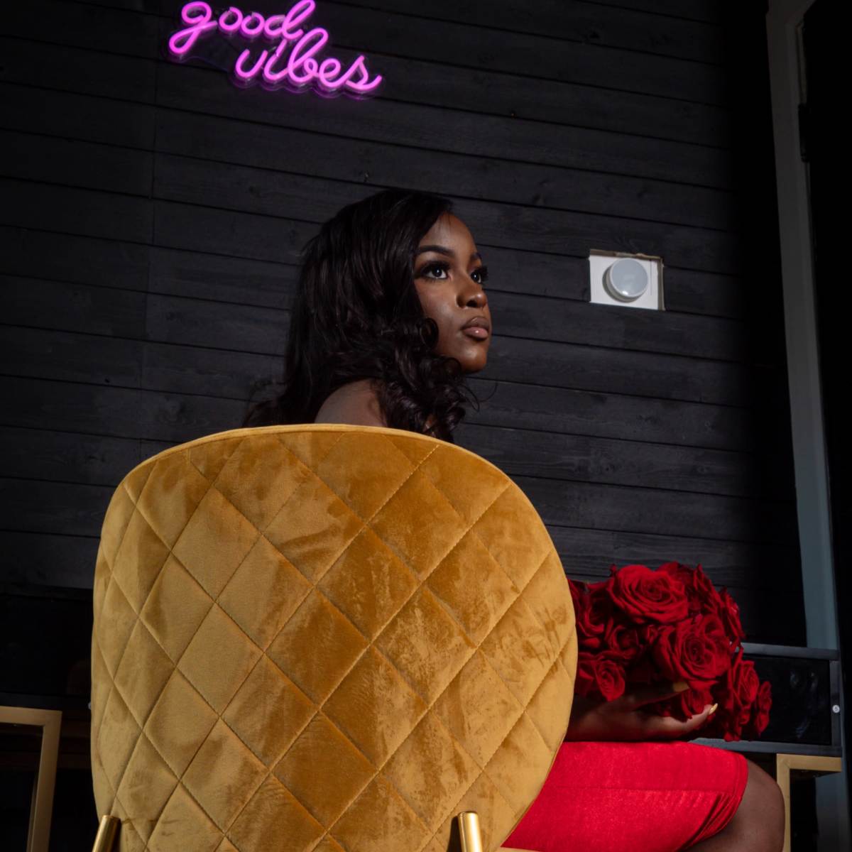 A woman holds a bouquet of red roses while seated on a mustard-colored chair in a dimly lit room with a neon sign that reads "good vibes."