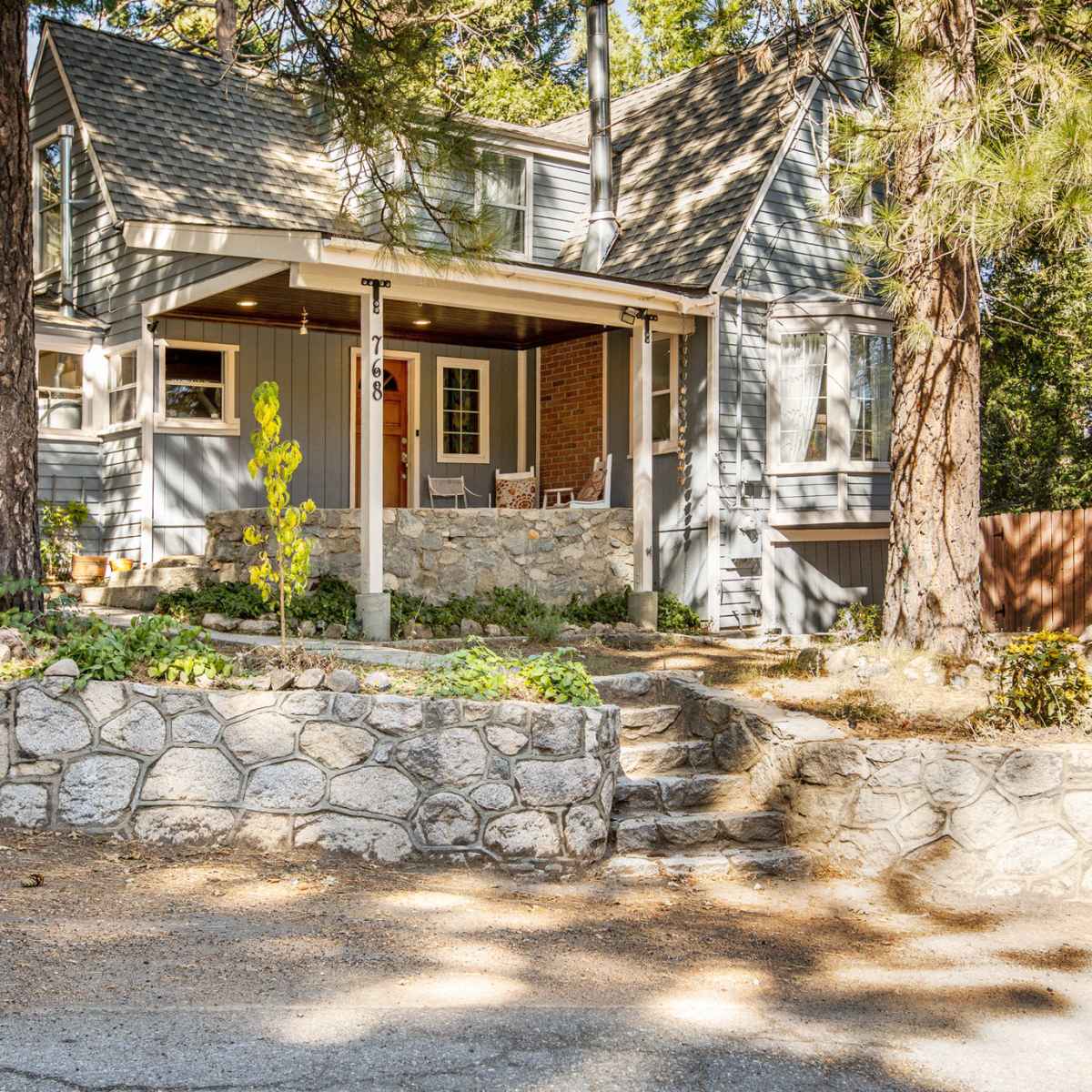 A two-story gray house with a stone porch and surrounded by tall trees and a landscaped yard is visible.