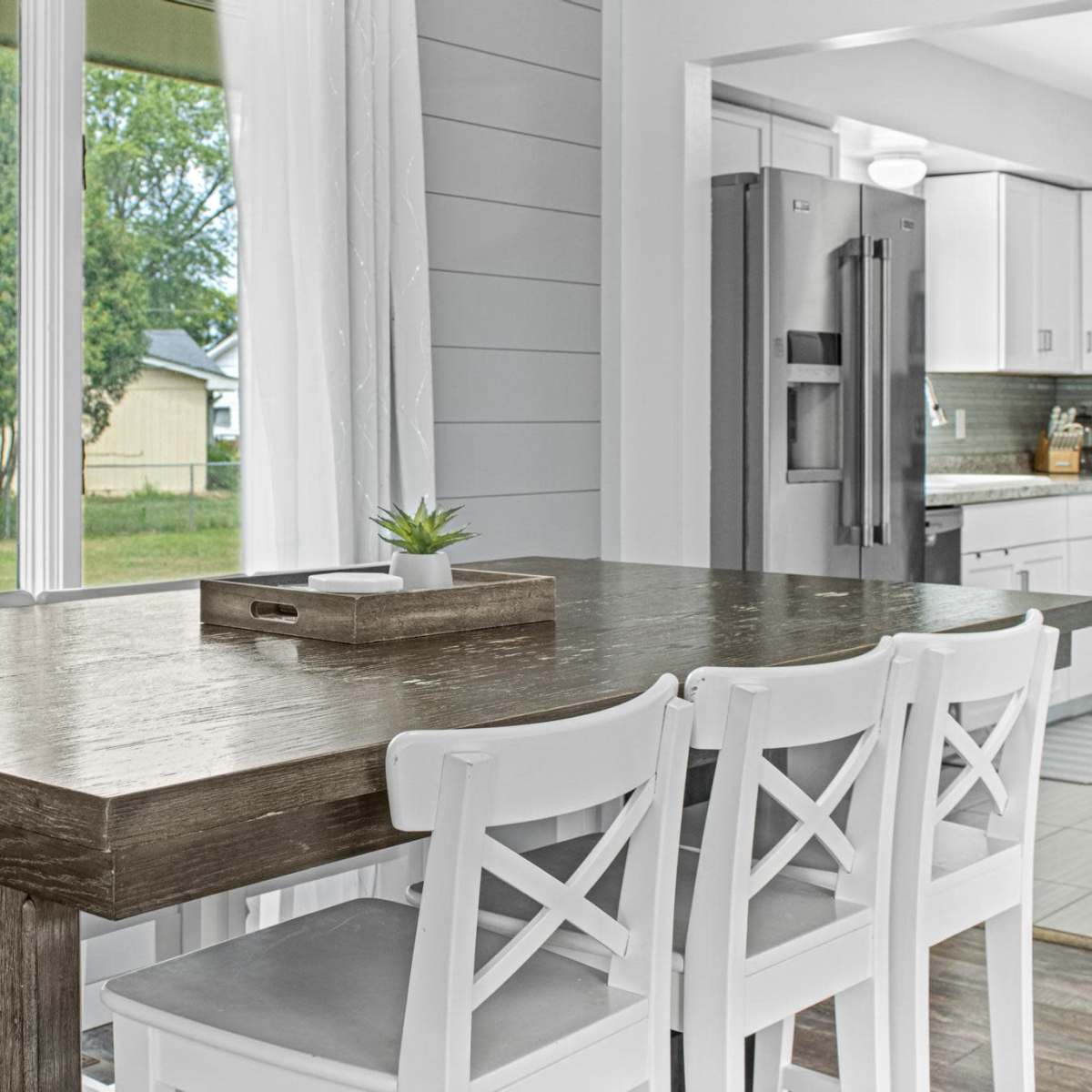 A wooden table with a tray sits in front of a large window, accompanied by white chairs in a bright kitchen with modern appliances.