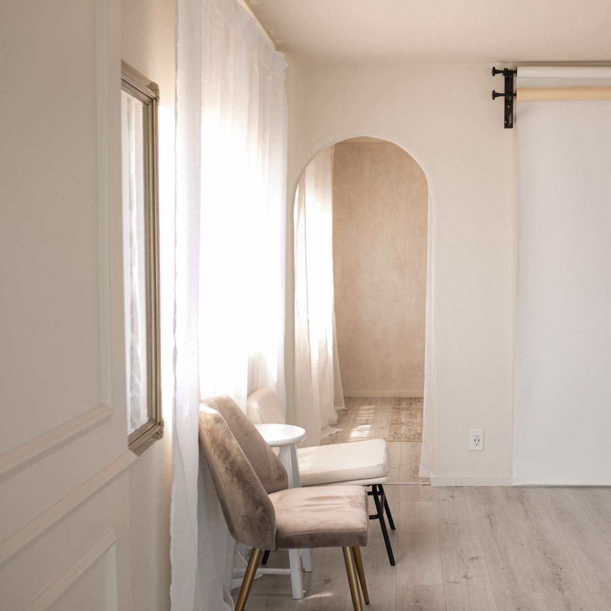 A minimalistic room featuring two chairs beside a small table, with an archway leading to a softly lit area and a blank backdrop on the wall.