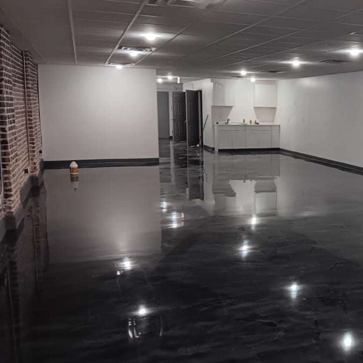 A large, empty interior space with a polished black floor and white walls, featuring a kitchenette in the background and a fire extinguisher mounted on the wall.