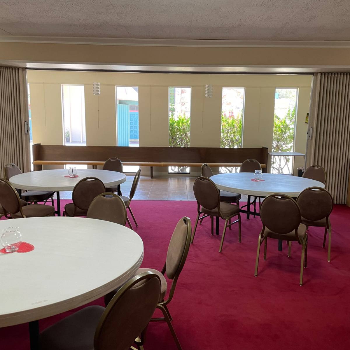 An interior room with several round tables and chairs arranged on a red carpet, featuring large windows that let in natural light.