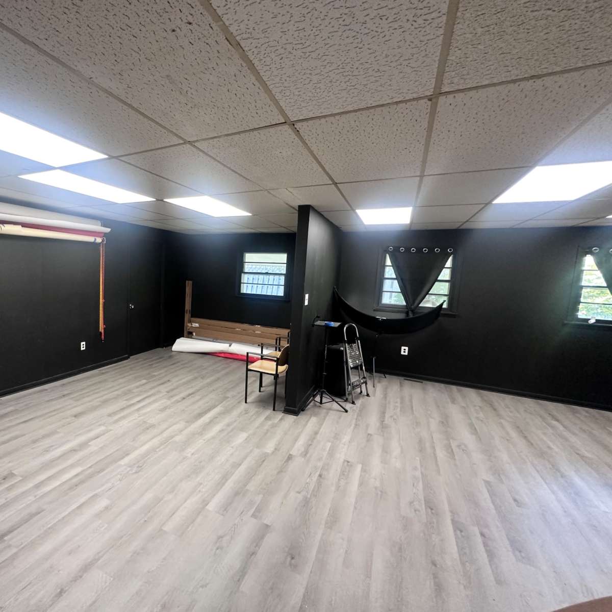 A spacious, empty room with black walls and large windows, featuring a chair and some rolled materials against one wall.