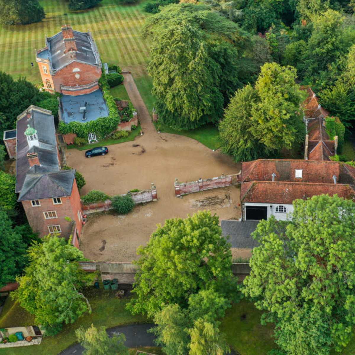 An aerial view shows a sprawling green landscape with several buildings, including a large house and a smaller structure, surrounded by dense trees and manicured lawns.