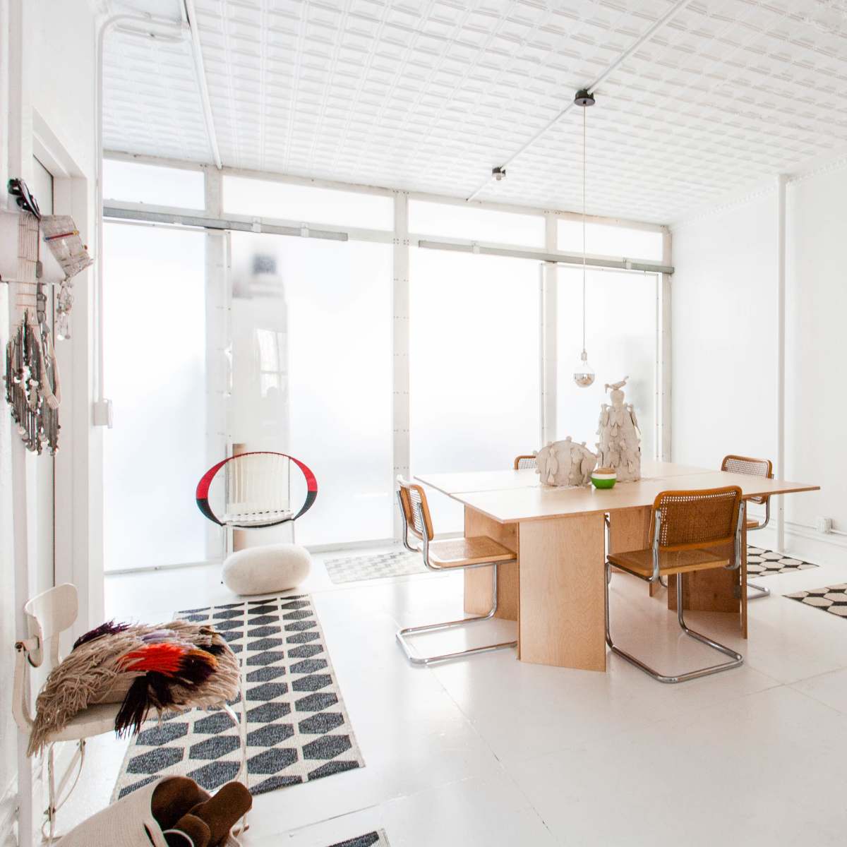 A minimalist dining area features a wooden table surrounded by metal-framed chairs, with patterned rugs beneath and large windows allowing natural light.