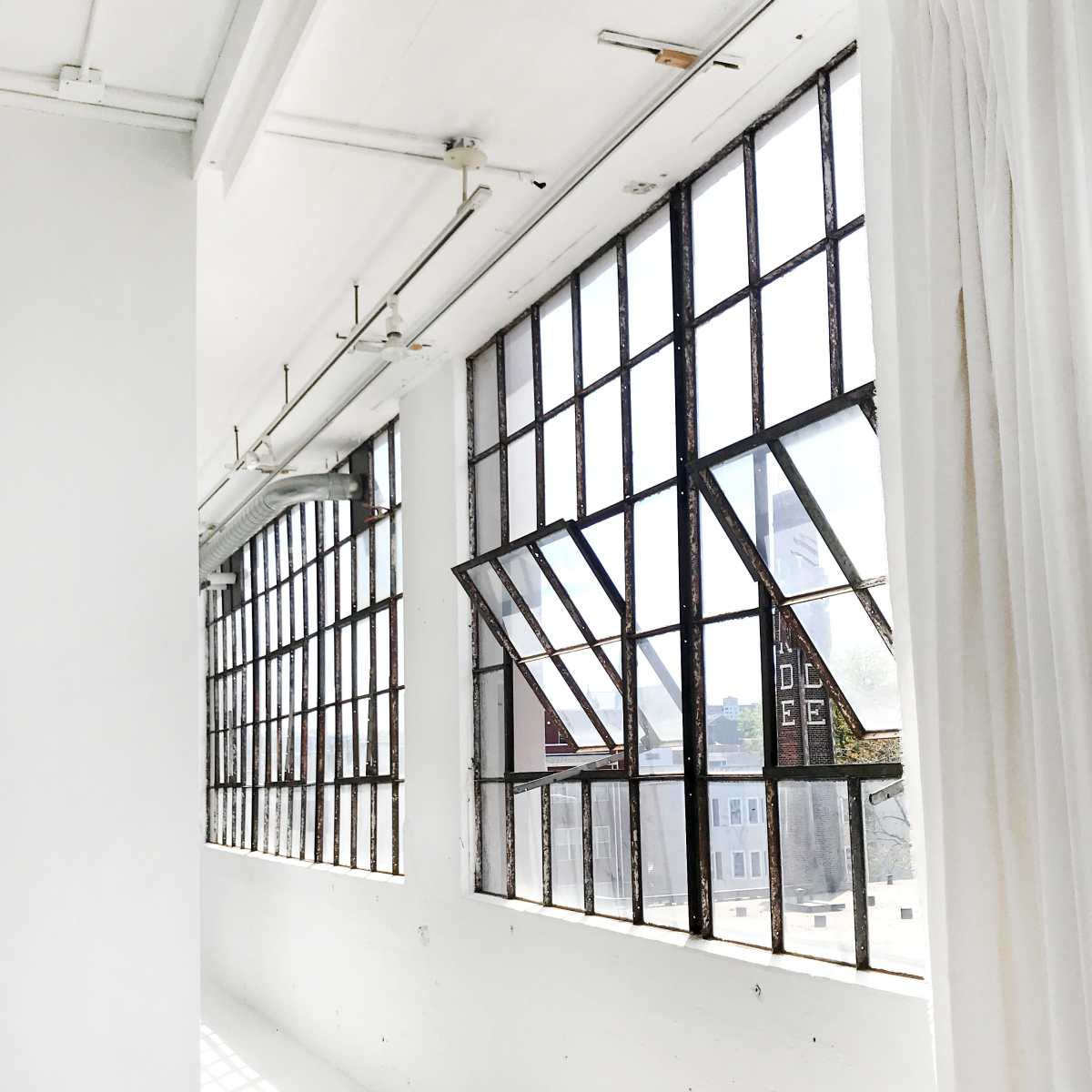 A bright, open room with large, industrial-style windows that are partially opened, allowing natural light to illuminate the white walls and floor.
