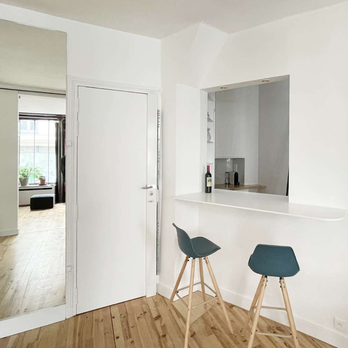 A minimalist interior space featuring a small bar area with two stools, a white wall, and a door leading to another room.