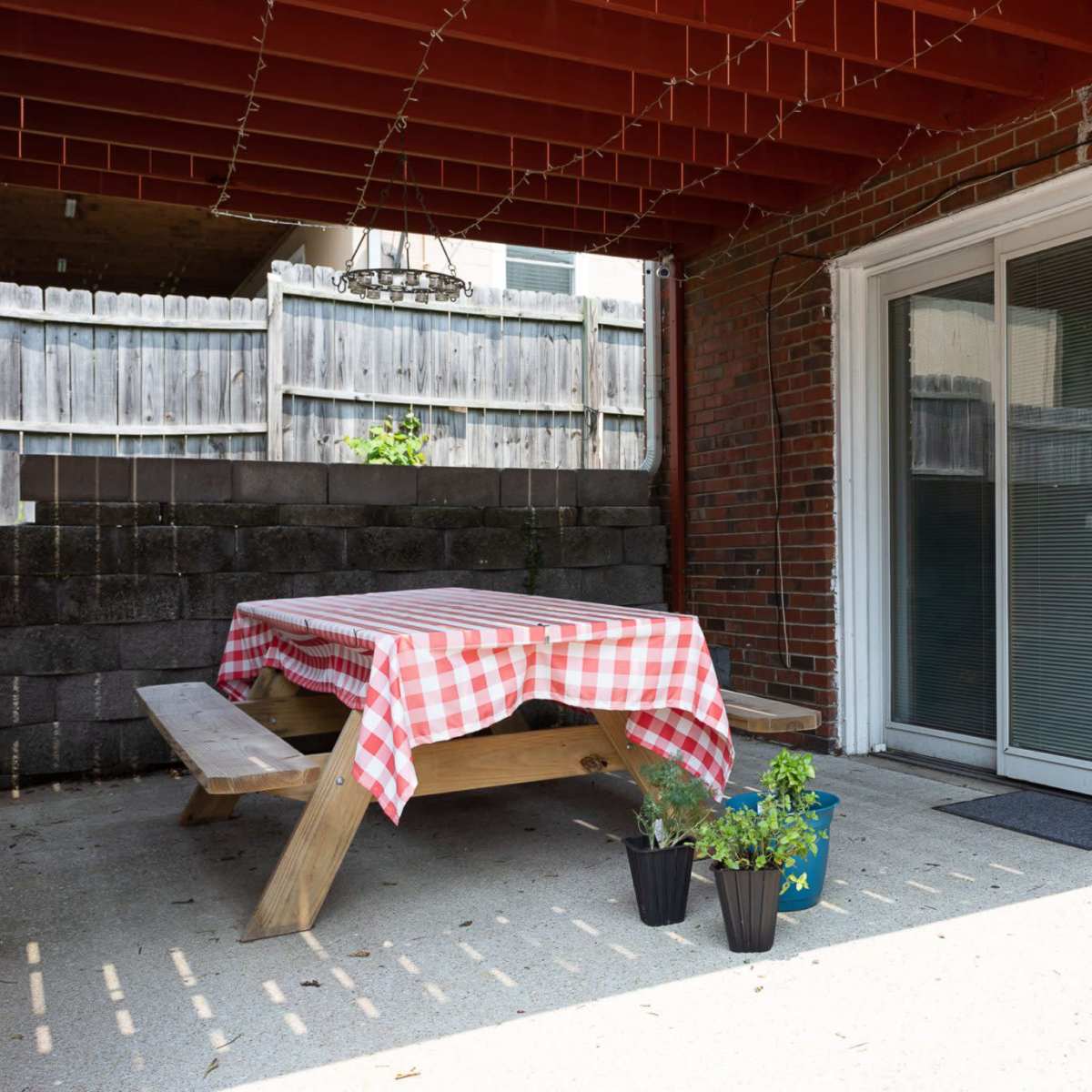 A covered patio features a wooden picnic table with a checkered tablecloth, flanked by two potted plants and sliding glass doors.