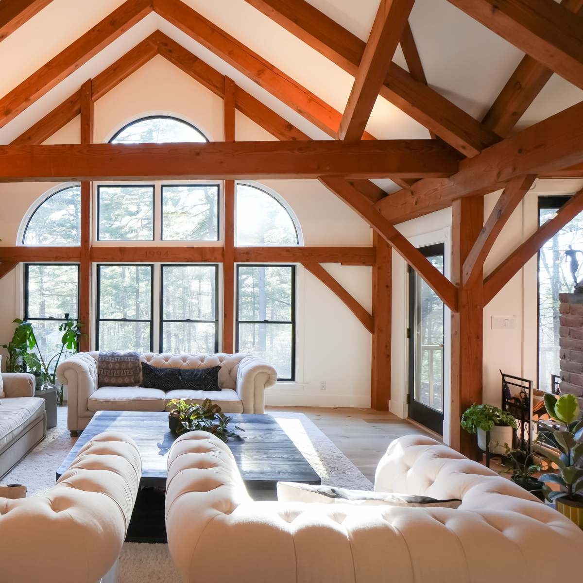 A spacious living room with a high, wooden-beamed ceiling, featuring two distinct sofas and large windows that provide natural light and a view of the outdoors.