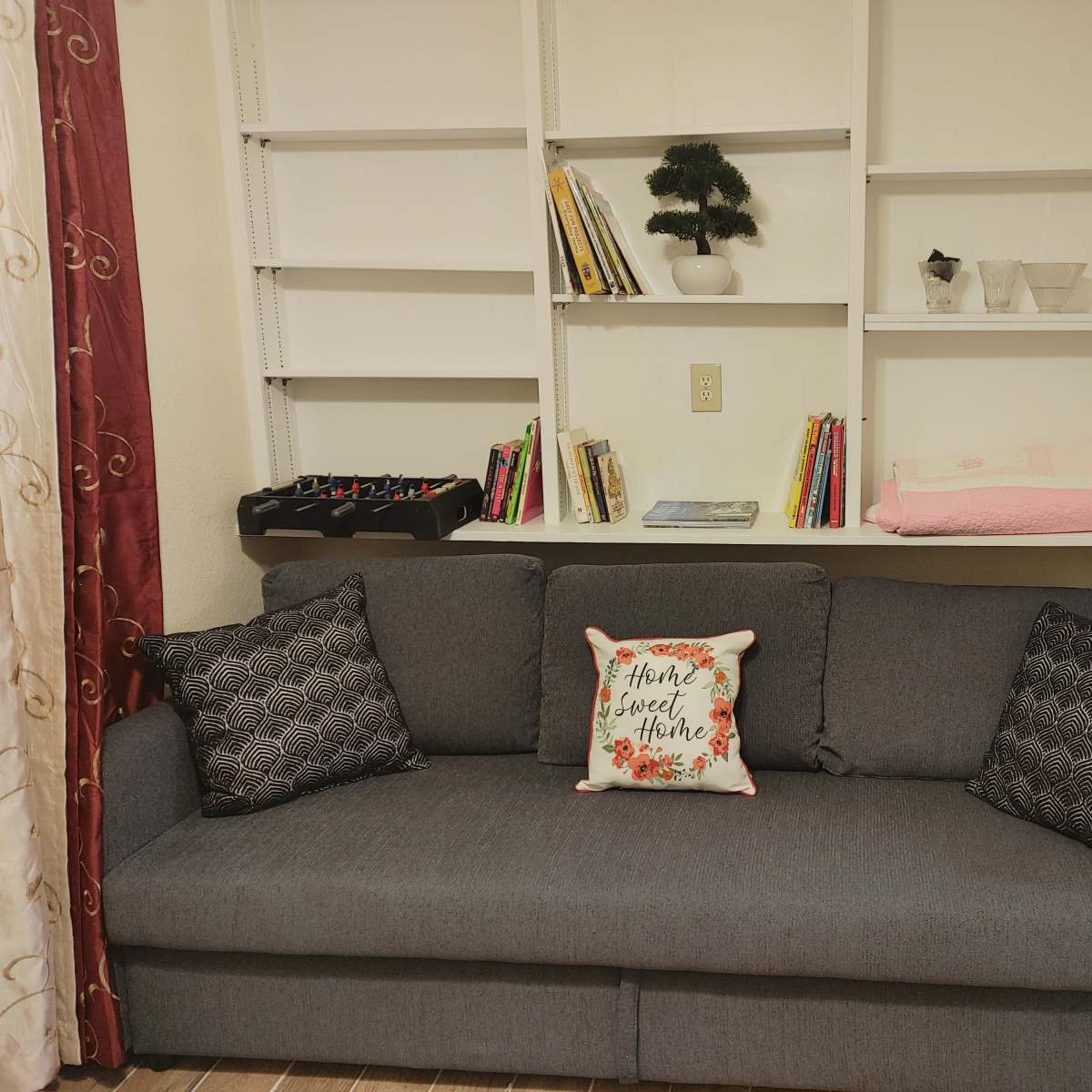 A gray couch with patterned pillows positioned in front of a wall-mounted shelf displaying books and decorative items.