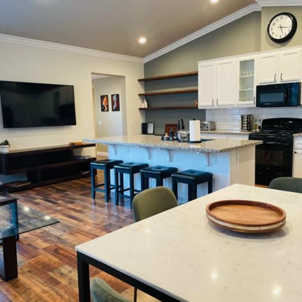 A modern kitchen and living area featuring a granite island with bar stools, a television mounted on the wall, and a dining table with a tray on it.
