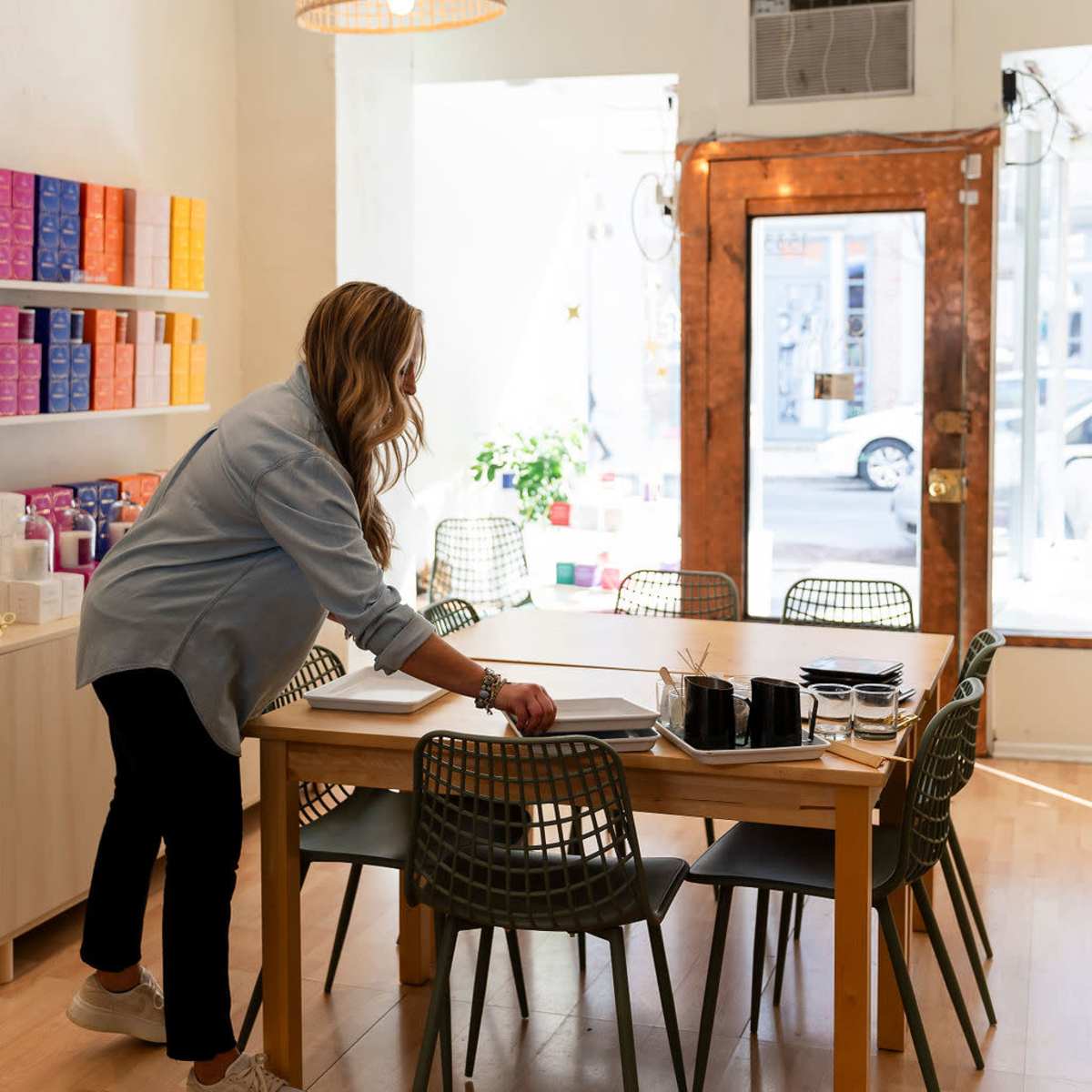 A woman adjusts items on a dining table in a brightly lit room with modern decor and a large window.