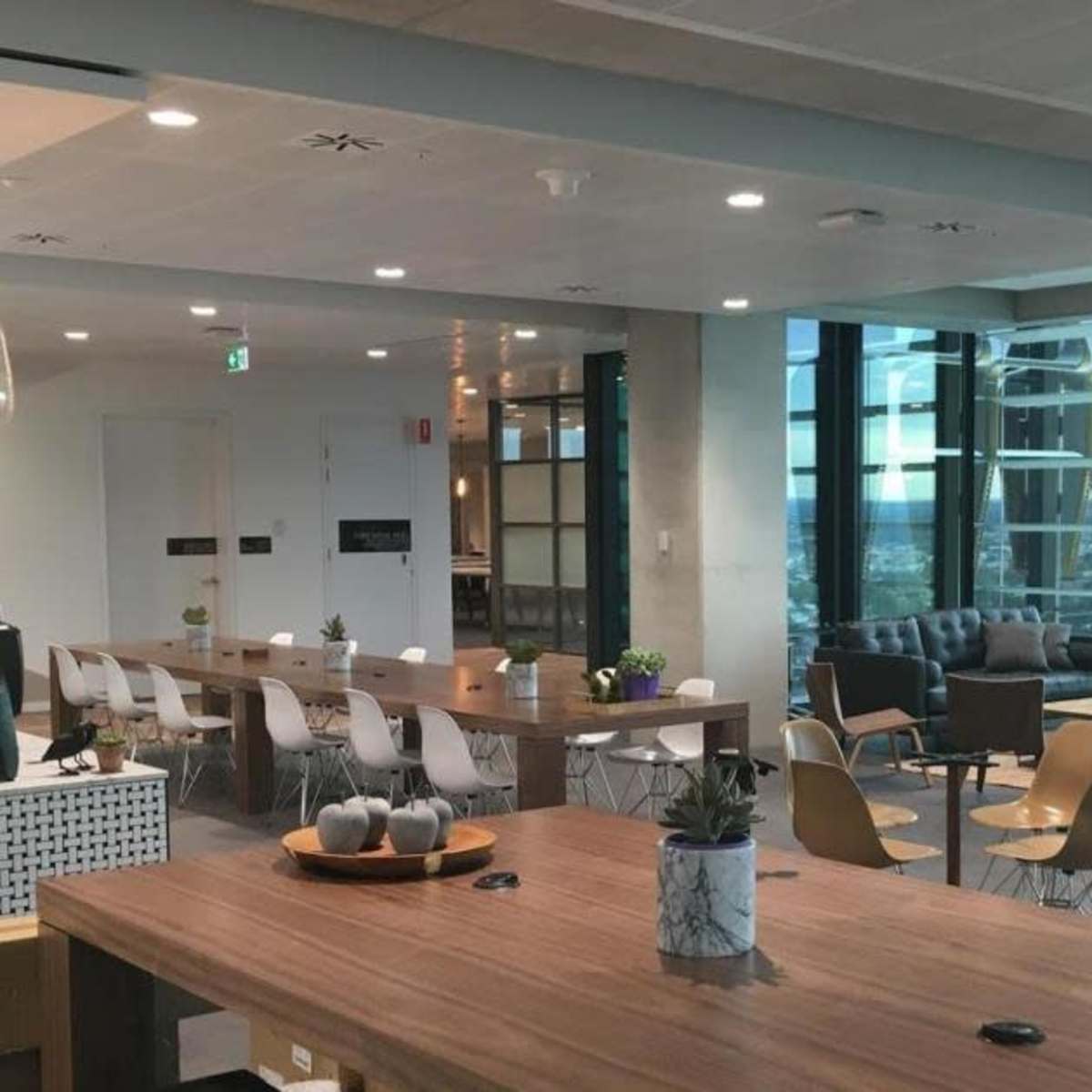 A modern, open-concept office space featuring a central wooden table, a coffee station, and various seating areas with large windows allowing natural light.