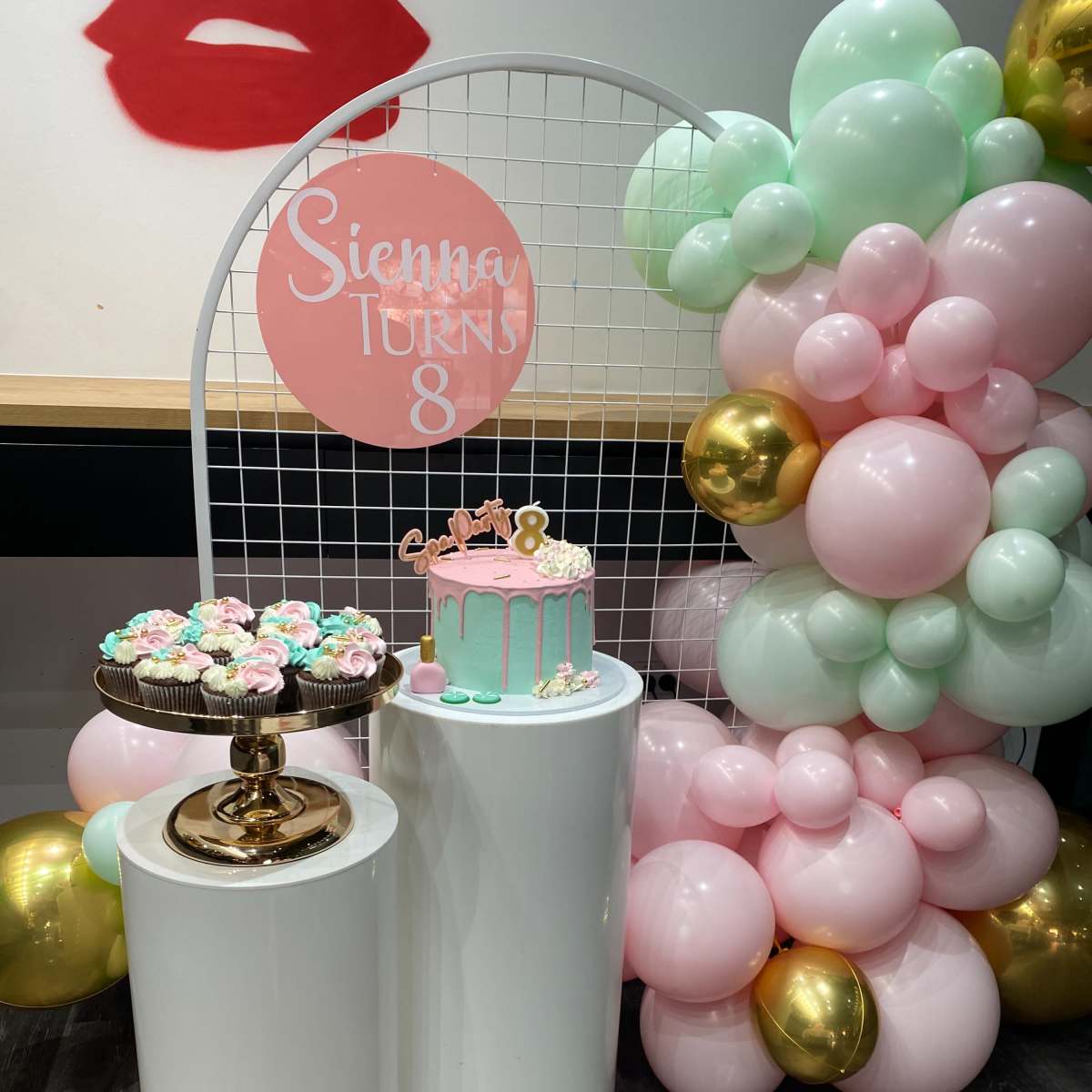A birthday celebration setup with a pink and mint green balloon backdrop, a cake on a pedestal labeled "Sienna Turns 8," and a decorative stand of cupcakes.