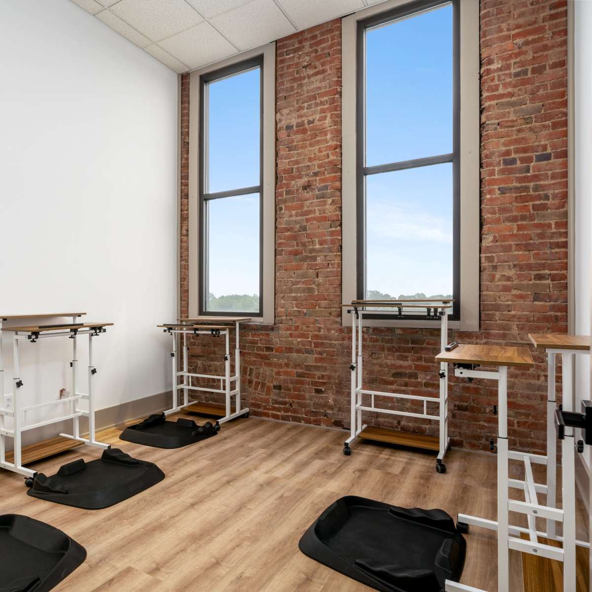 A room with wooden flooring, exposed brick walls, and multiple adjustable standing desks positioned near large windows.