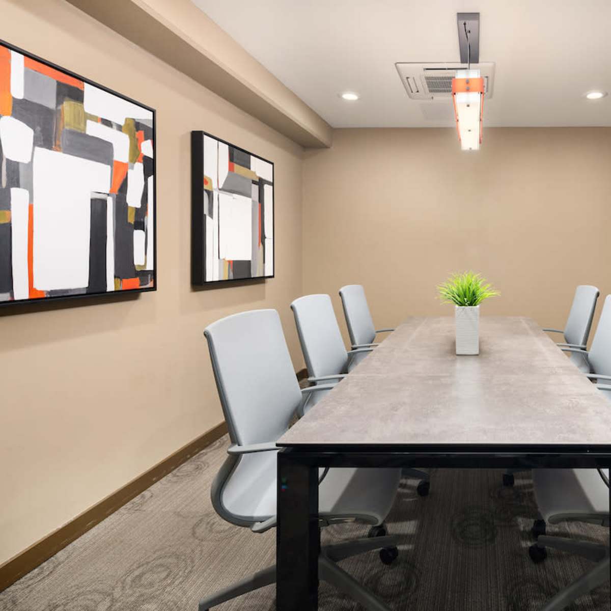 A modern conference room features a long table surrounded by six gray chairs, with artwork on the walls and a small plant centerpiece.