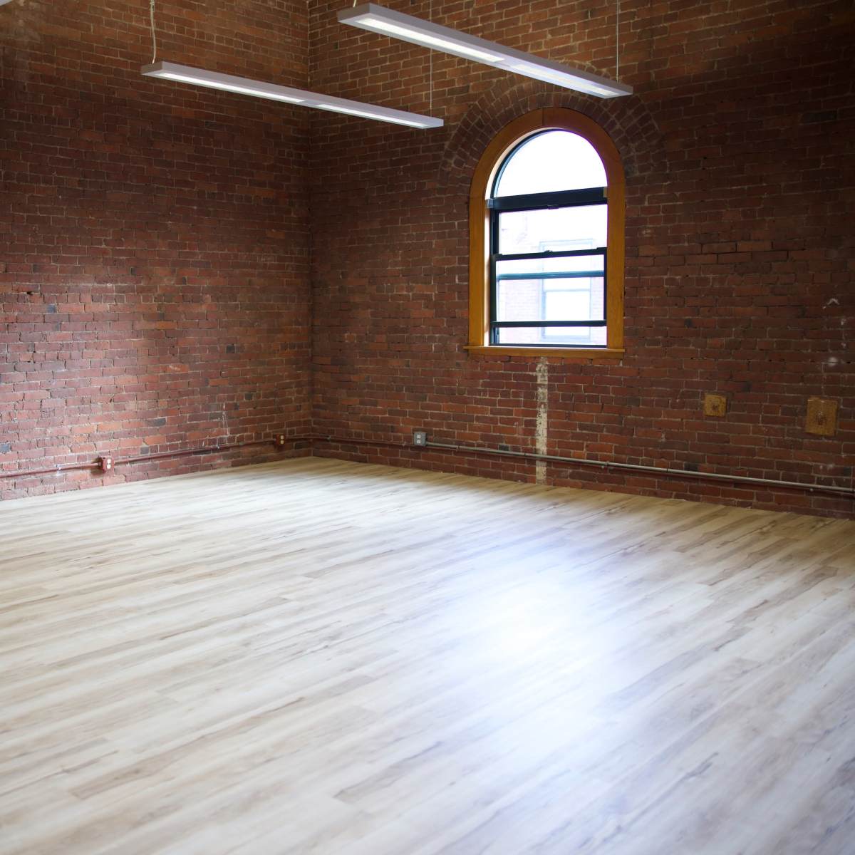 An empty room with exposed brick walls and a large window letting in natural light, featuring a wooden floor.