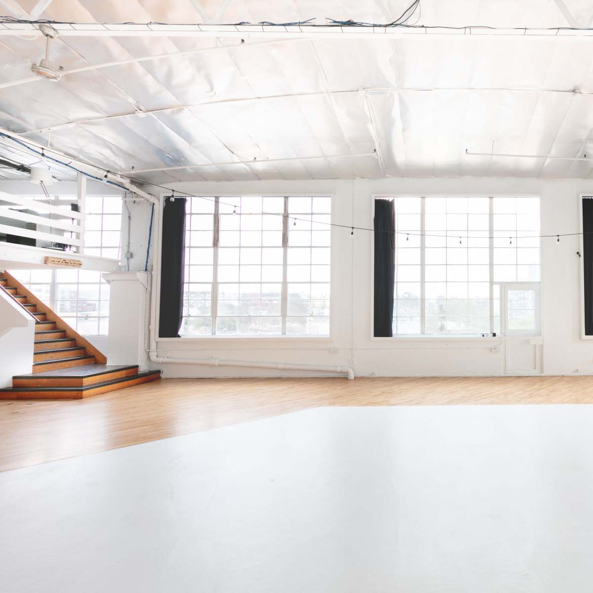 A spacious, brightly lit room with large windows, a wooden floor, a staircase, and a minimalist couch in the corner.