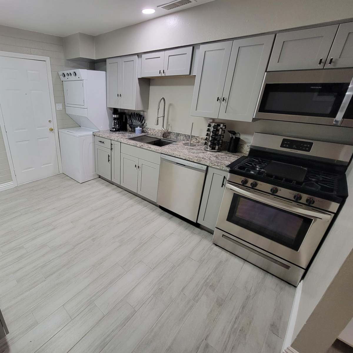 A modern kitchen featuring gray cabinets, stainless steel appliances, a granite countertop, and a laundry area with a stacked washer and dryer.