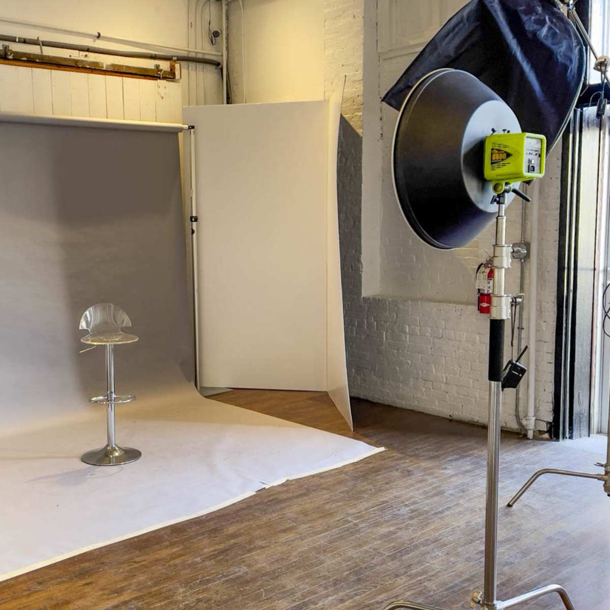 A photography studio setup with a gray backdrop, a white backdrop, a modern stool in the center, and a large lighting umbrella on a stand.