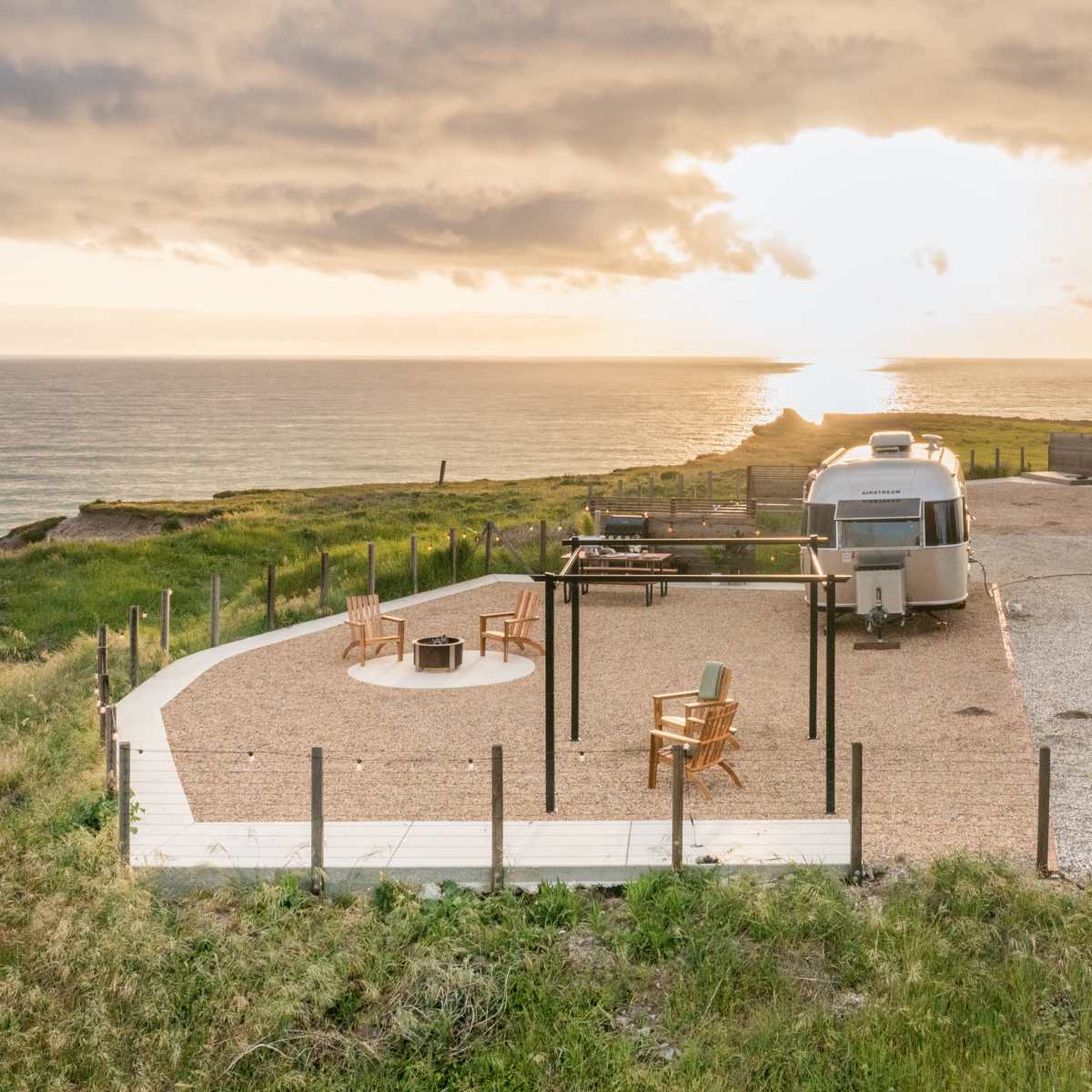 A trailer is parked on a gravel area overlooking the ocean, with wooden seating around a fire pit and a sunset in the background.