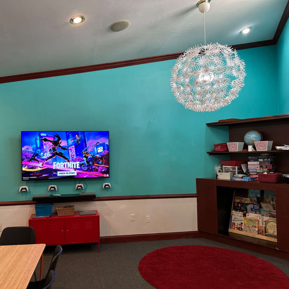 A colorful room with a television displaying a "Fortnite" screen, a circular rug on the floor, a bookshelf filled with books, and a modern light fixture hanging from the ceiling.