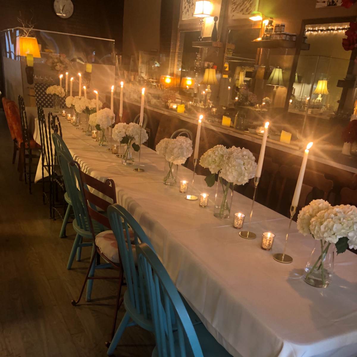 A long dining table is set with white flowers in glass vases and lit candles, surrounded by colorful chairs in a warmly lit restaurant.