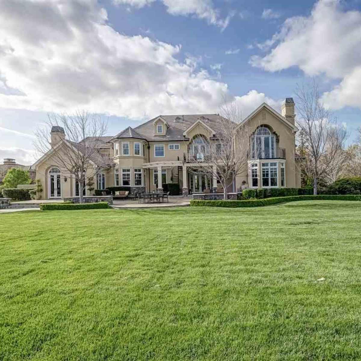 A large, two-story house with a manicured lawn and several trees sits under a partly cloudy sky.