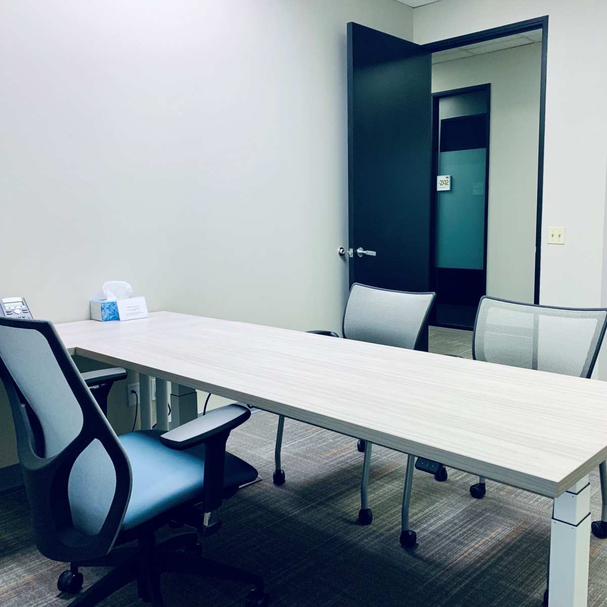A small office with a long wooden table, three chairs, a phone, and an open door leading to another room.