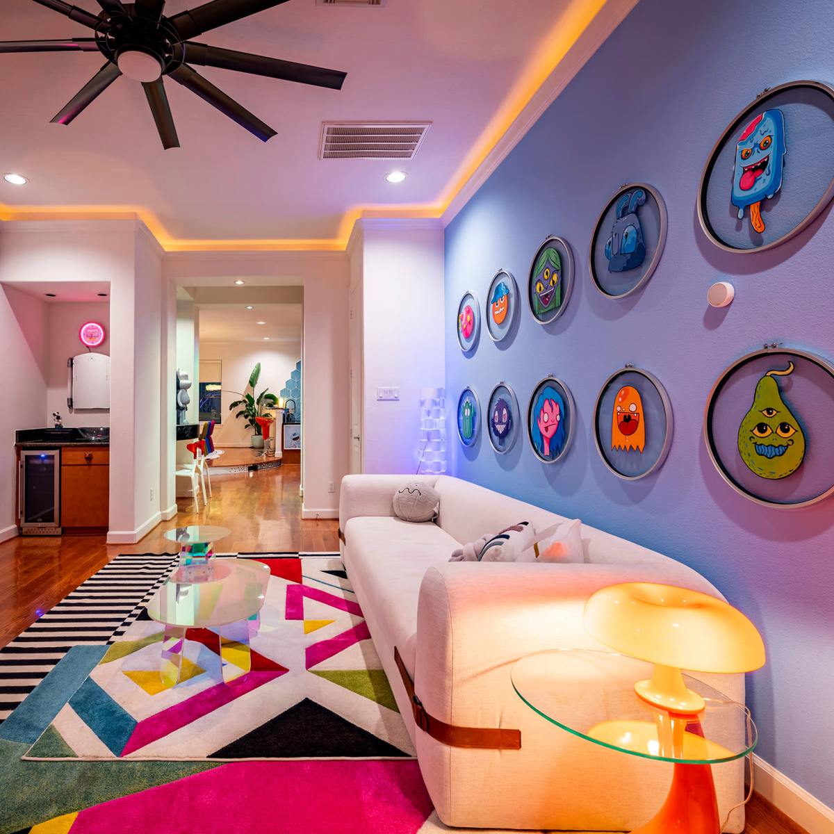 A modern living room with a white sofa, colorful patterned rug, wall-mounted artwork, and a kitchen area visible in the background.