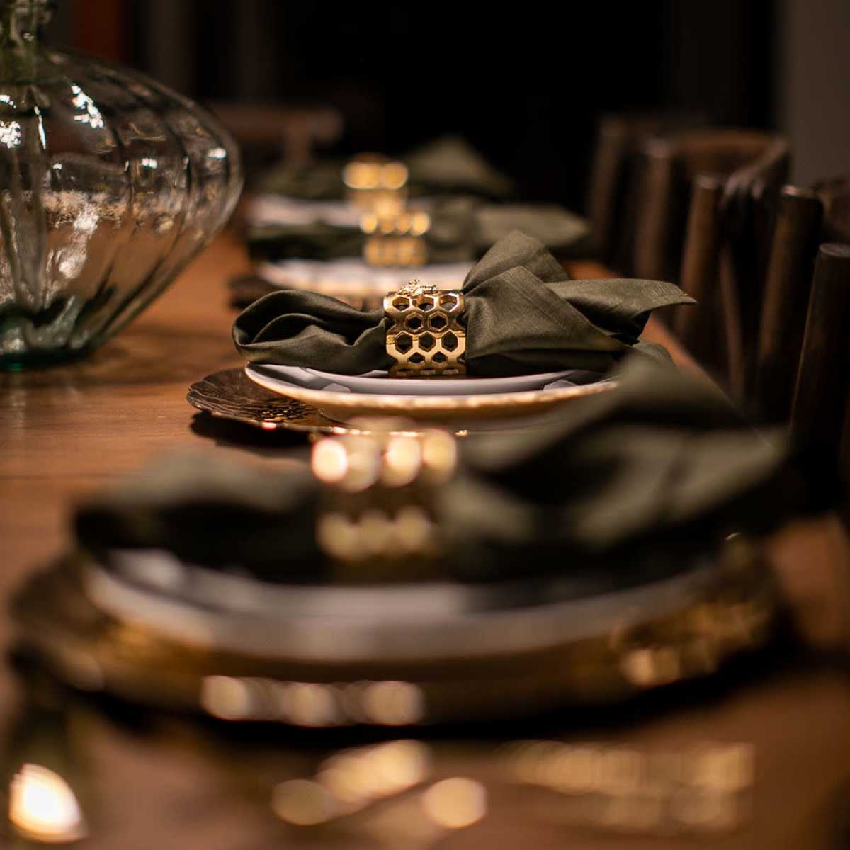 A wooden dining table is elegantly set with plates, green napkins, and gold napkin rings, complemented by a decorative vase of greenery.