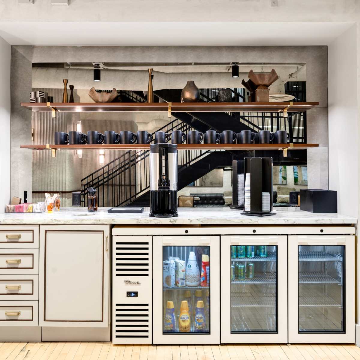A modern kitchen bar area features a marble countertop with open shelves above and a glass-front refrigerator below, displaying beverages.